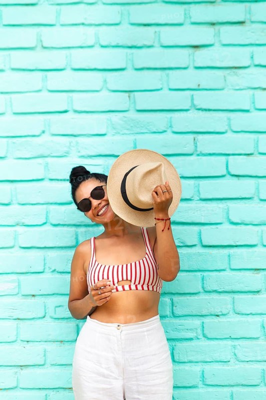 A young Latin woman in sunglasses and summer attire, smiling and holding a hat against a vibrant turquoise brick wall in Mancora, Peru, capturing a sunny, joyful moment