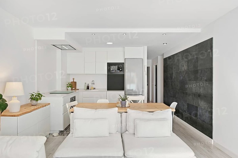 A chic, open concept kitchen and living area features sleek white cabinets, a natural wood table, and a black slate accent wall, creating a modern minimalist aesthetic in the city center