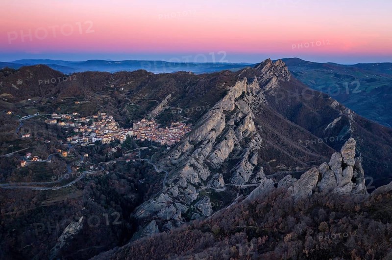 A stunning aerial view of Castelmezzano at sunrise, nestled in the rugged Lucanian Dolomites. The village glows softly under the pink and purple hues of dawn, surrounded by dramatic cliffs