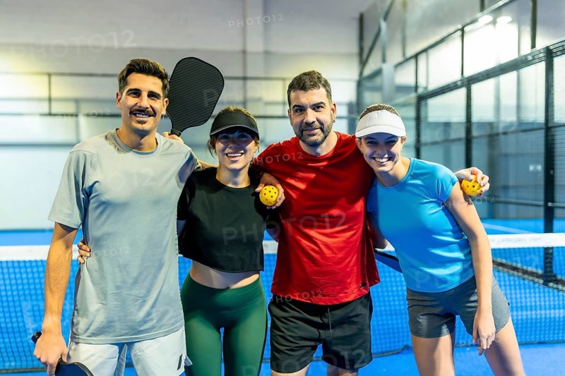 Four smiling friends hugging on a blue pickleball court, holding paddles and balls, celebrating friendship, teamwork and active, healthy recreation together