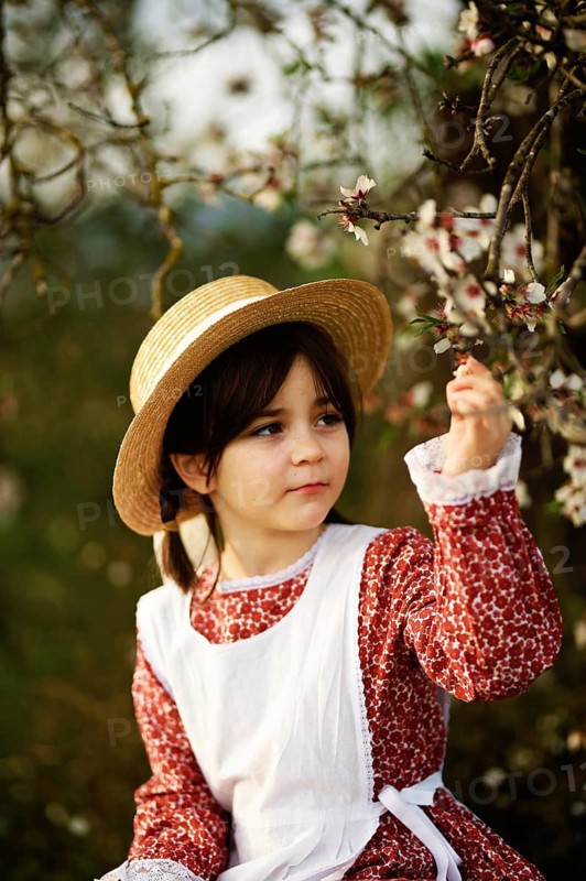 A child wearing a straw hat and vintage dress observes the blossoms on an almond tree branch. This serene scene captures the essence of spring and childlike curiosity in the midst of nature