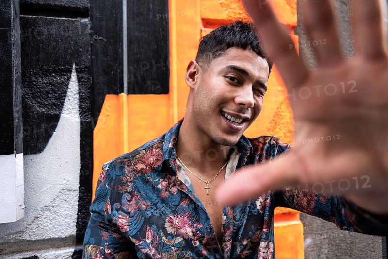 A cheerful man in a floral shirt playfully poses with his hand extended towards the camera against a vibrant graffiti wall, exuding energy and joy