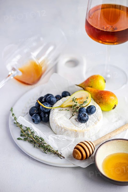 A delectable cheese and fruit platter featuring creamy brie cheese, ripe pears, fresh blueberries, and aromatic herbs, elegantly arranged with honey and a glass of amber wine on a neutral backdrop