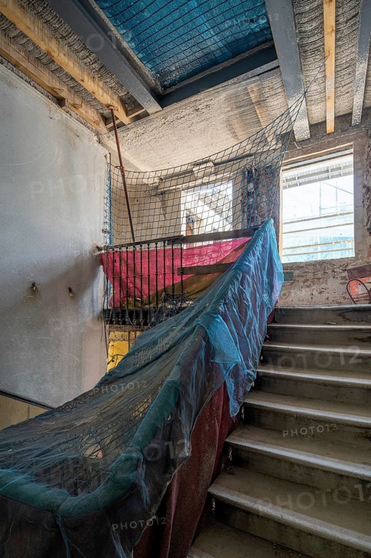 Sunlit construction site with stairs, partially covered with protective nets. Exposed beams and renovation materials create a dynamic, unfinished atmosphere