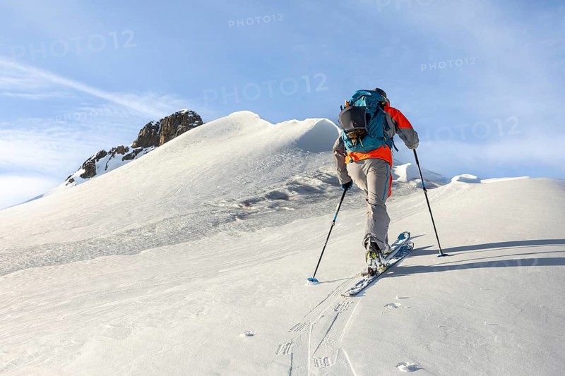 A skier with a backpack and poles ascends a snowy mountain, epitomizing the adventure and challenge of ski touring against a backdrop of clear skies and rugged terrain