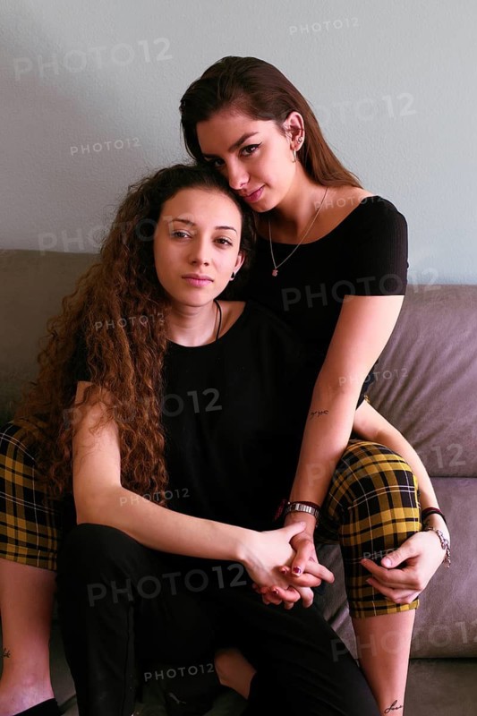 A tender portrait of a female gay couple embracing on a sofa. Both women gaze lovingly into the camera, highlighting their connection and affection
