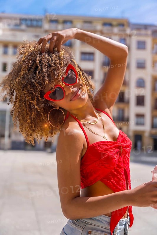 Vertical photo of a lovely latin young woman in summer clothes and funny red heart shape sunglasses posing in the city