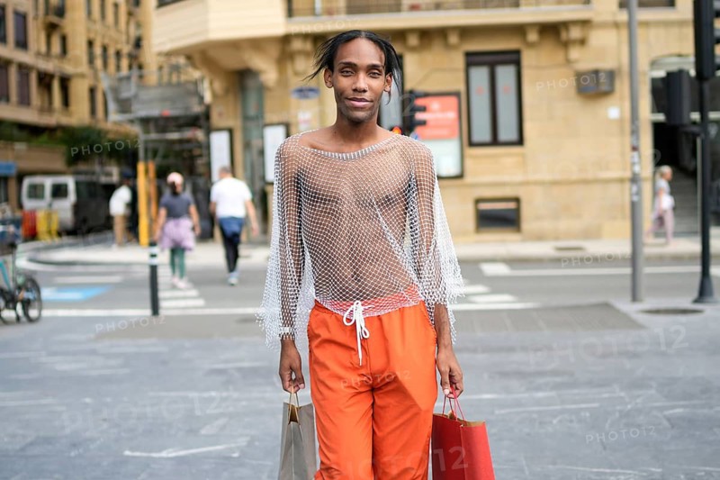 A man walks confidently in the city, showcasing vibrant fashion with orange pants and a mesh top. He carries shopping bags, embodying pride and an expressive lifestyle