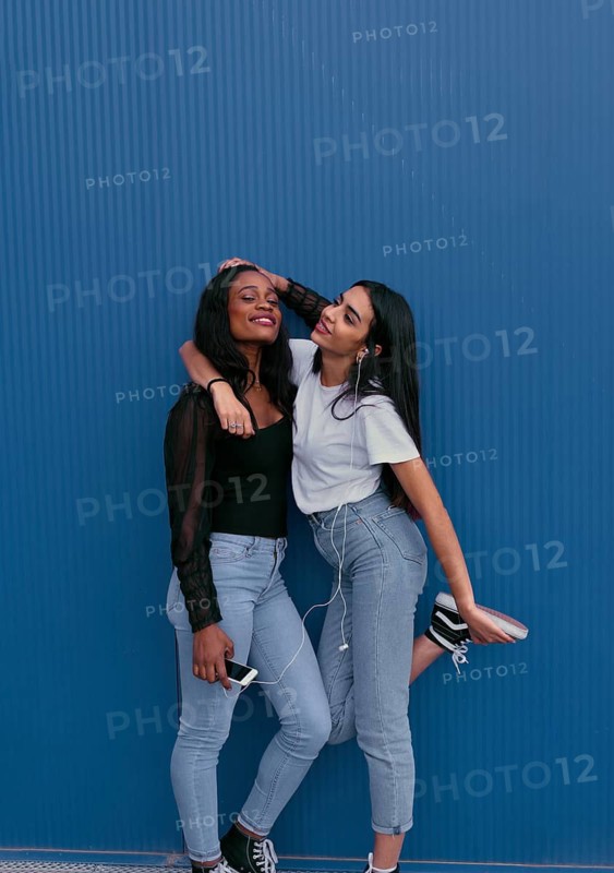 Two young friends stand against a blue wall, sharing headphones and enjoying music. They are smiling, expressing joy and friendship, dressed casually, conveying a relaxed vibe