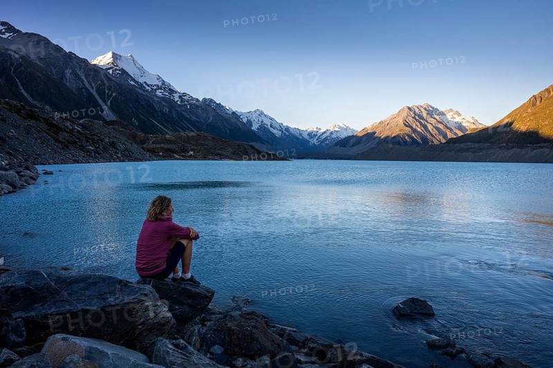 A woman is sitting on a rock at Tasman Lake. On the left, Mount Cook. Golden hour. Aoraki Mount Cook National Park, Canterbury, South Island, New Zealand
