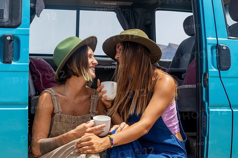 Two lesbian women share a joyful moment outside a blue van, sipping coffee under the sun. They wear matching hats, showcasing a love for travel and adventure