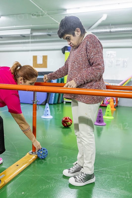 A therapist assists a person with physical therapy exercises in a mental health residence, focusing on balance and coordination. The environment is supportive and therapeutic
