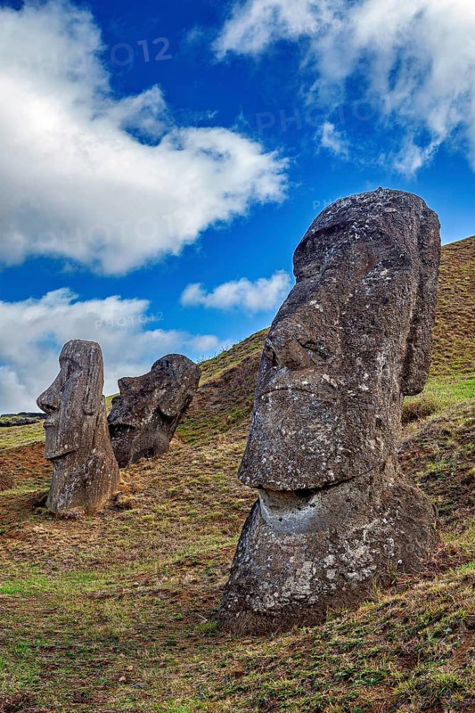 Large stone sculptures on a grassy hill under a cloudy blue sky, The Moai Statues on Easter Island in the Pacific Ocean