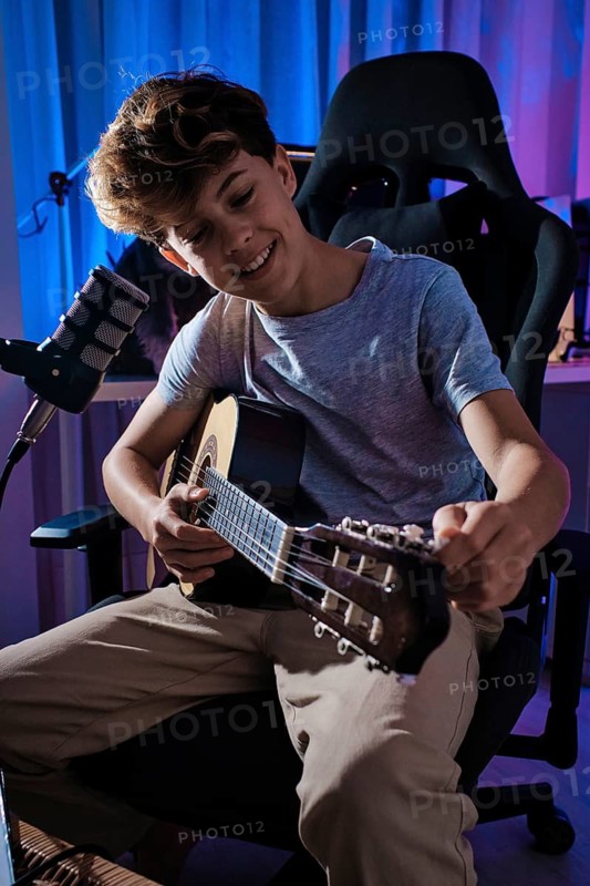A young boy is tuning a guitar while sitting in a comfy chair, surrounded by colorful lighting. A microphone is positioned nearby, showcasing a creative atmosphere at home