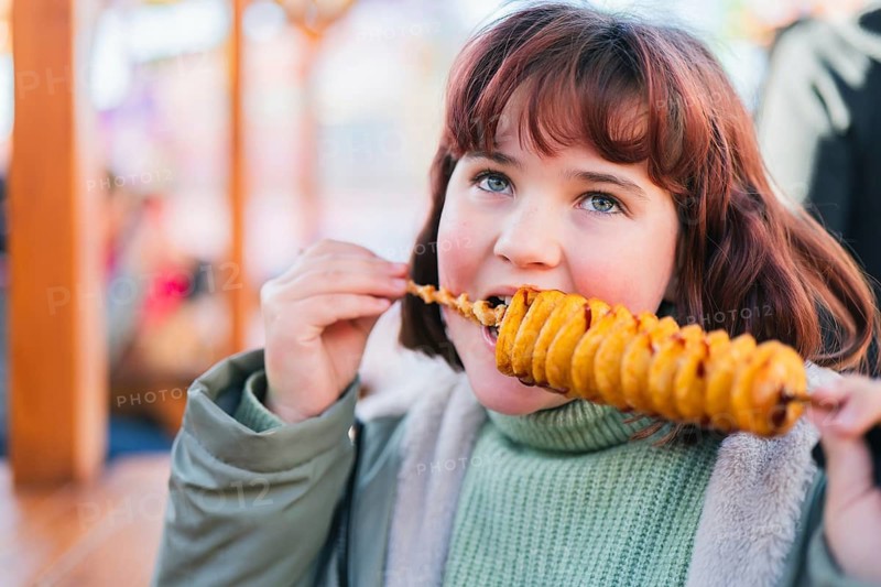 A young girl enjoys a spiral tornado potato on a stick at an outdoor market. The golden, crispy snack highlights the festive atmosphere and brings joy and excitement