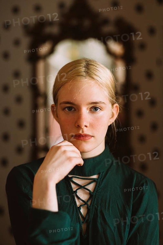 A poised young woman with blonde hair and blue eyes wears a dark green lace-up dress, gazing confidently at the camera. A vintage mirror and soft lighting add a classic, timeless feel
