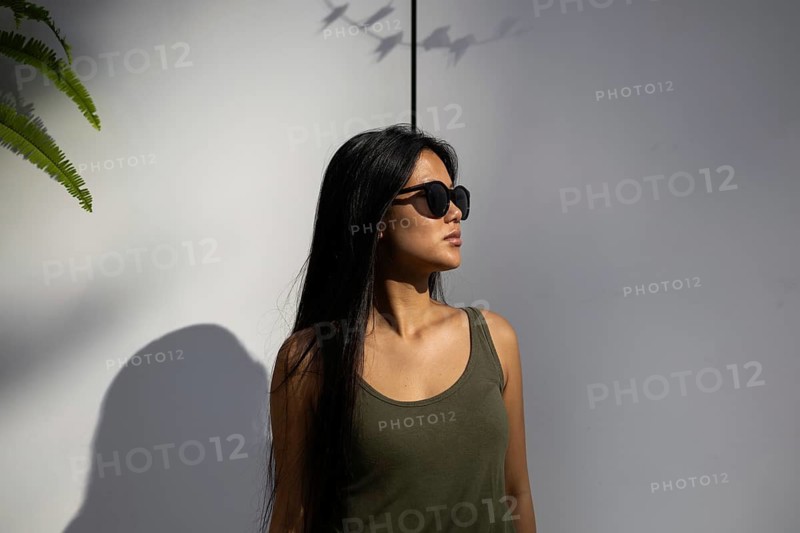 Chinese woman in sunglasses standing against a sunlit wall, basking in the summer glow, embodying urban style and confidence in a vibrant city setting