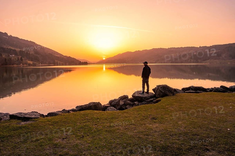 A person looks at a lake at sunset surrounded by hills and rocks, Großer Alpsee, Immenstadt im Allgäu, Bavaria, Germany