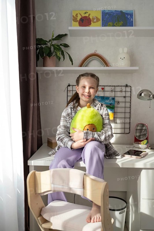 A cheerful girl sits in a sunlit room, surrounded by plush toys and colorful decor. The cozy space includes a fun and whimsical atmosphere that appeals to all ages
