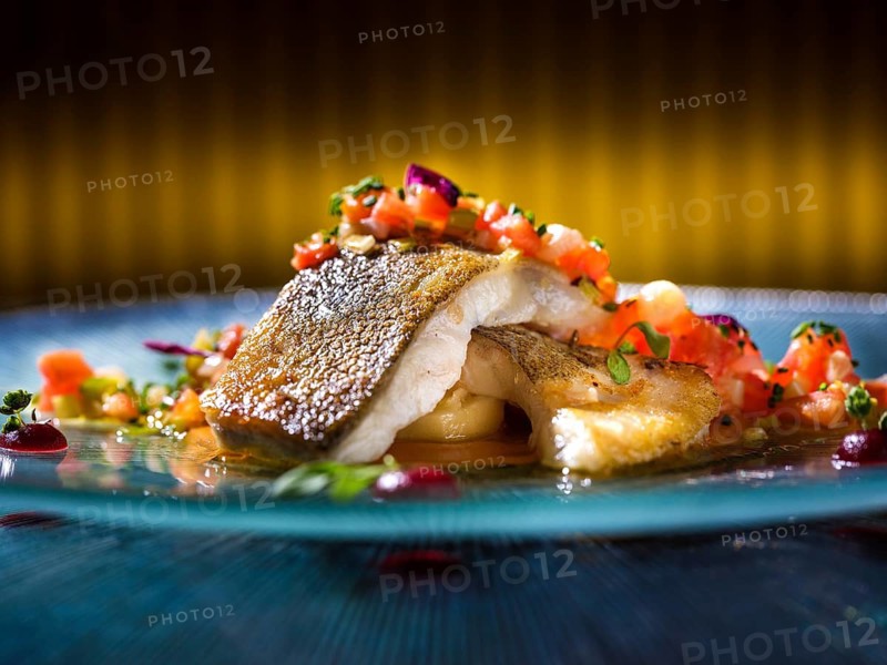 A beautifully plated gourmet hake fillet is garnished with vibrant diced tomatoes, fresh herbs, and delicate vegetable garnish, set against a soft focused background