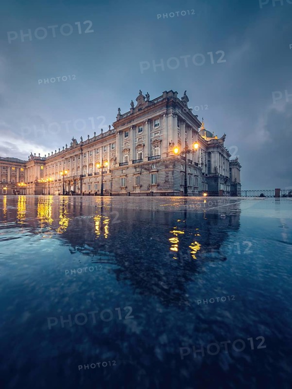 Majestic Royal Palace of Madrid with illuminated facade in twilight. The architecture is mirrored in a rain soaked surface, creating a serene, reflective scene