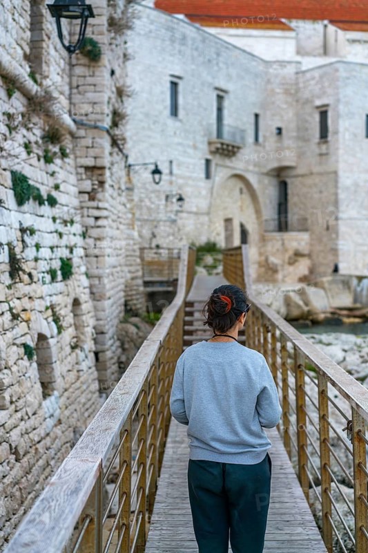 A person walks along a wooden bridge leading to the historic stone architecture of Polignano a Mare, a picturesque castle village on the stunning Puglia coastline