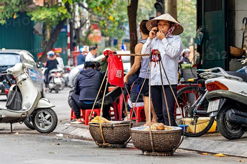 Street vendor with straw hat talking on the phone in a busy street with scooters in the background, A woman talking on the phone in the streets of Hanoi Vietnam