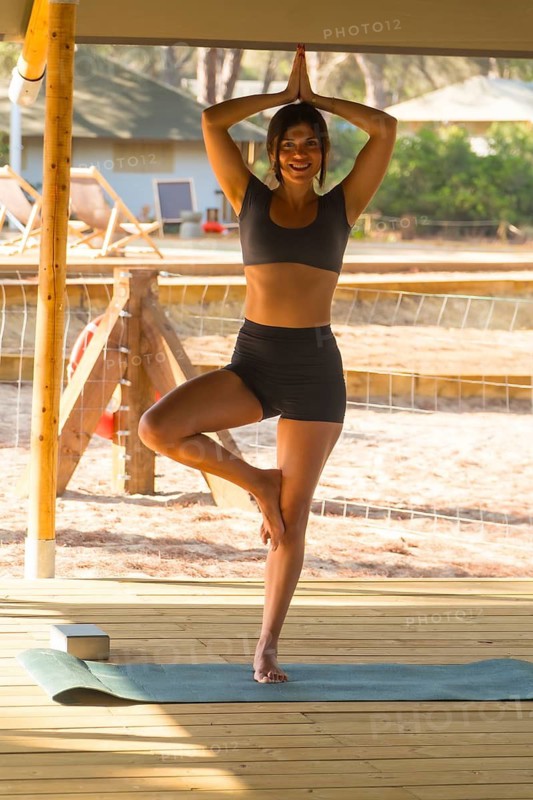 Young woman practicing yoga in tree pose, showcasing balance and mindfulness on a wooden deck surrounded by serene nature, embodying health and wellness in a tranquil outdoor environment