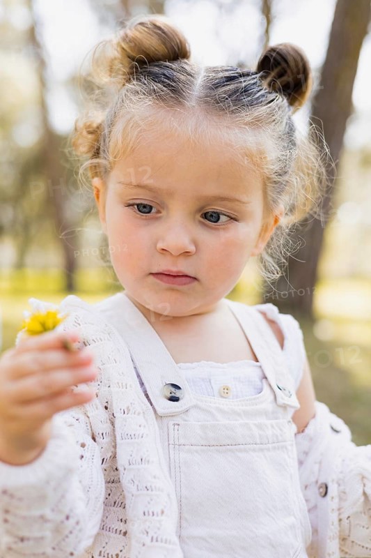 A young girl stands outdoors in a sunlit park, holding a small yellow flower. She wears a white outfit and looks attentively at the bloom, surrounded by trees and nature