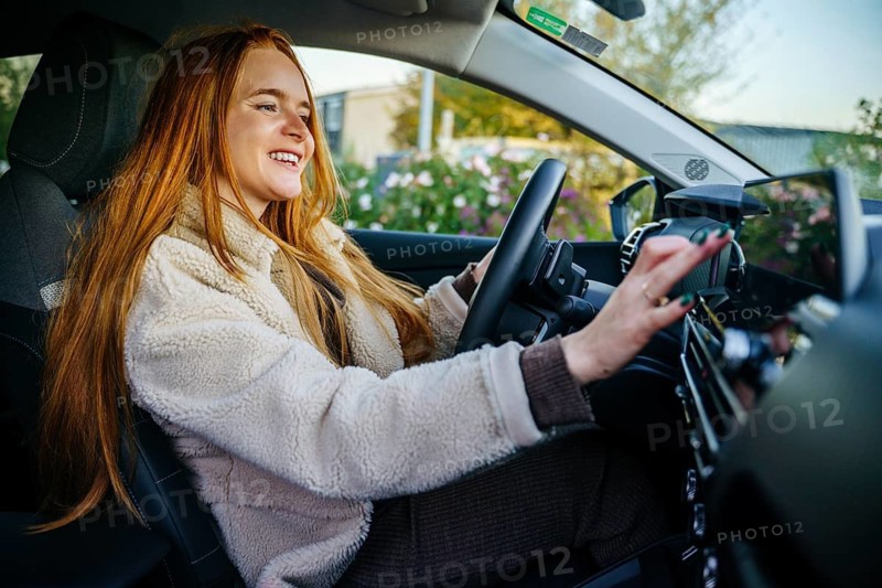 A beautiful pregnant woman with long red hair drives a car, enjoying a sunny day. Her smile radiates happiness, showcasing an uplifting moment of freedom and joy