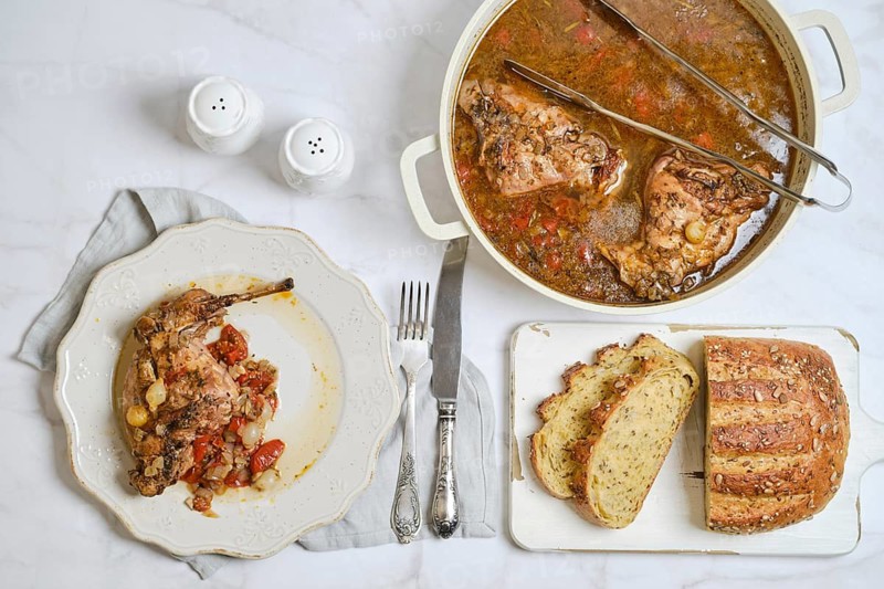 A plate with rabbit stew with vegetables next to a pot filled with more stew. Slices of bread lay on a wooden board. Salt and pepper shakers sit on a clean tablecloth