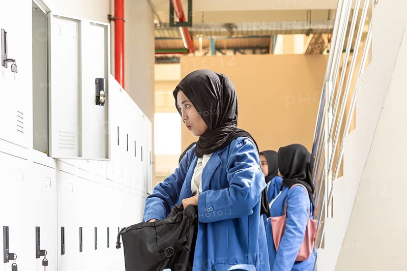 Asian Muslim student in a school hallway, wearing a blue uniform, organizing her belongings in a locker. She is preparing for a day of learning with a focused expression