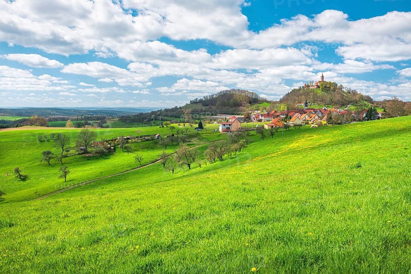 View of hilly landscape with the village of Seitenroda and Leuchtenburg in spring, in front a green meadow with yellow dandelion blossoms, near Kahla, Thuringia, Germany