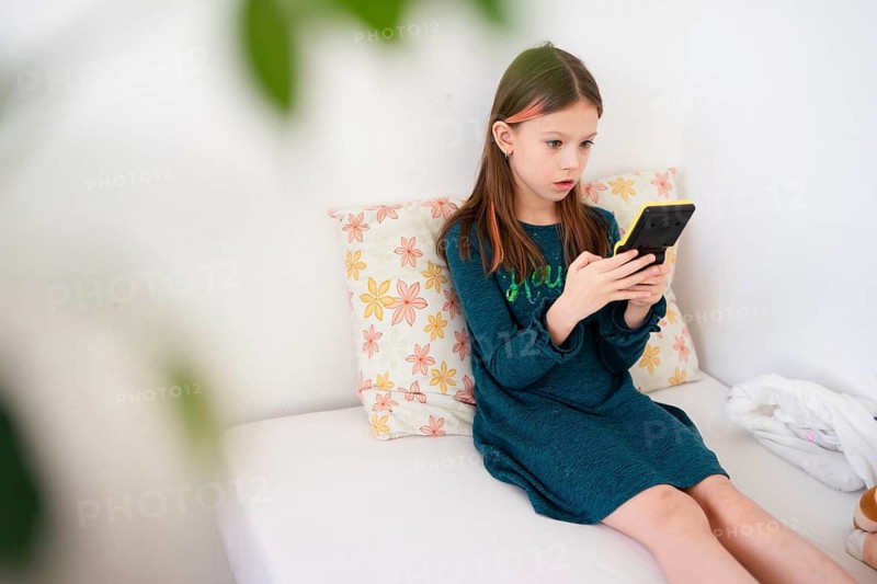 A young girl in a teal dress sits on a white bed, deeply focused on her handheld gaming console. The cozy indoor setting, soft lighting, and floral pillow create a relaxed and immersive atmosphere