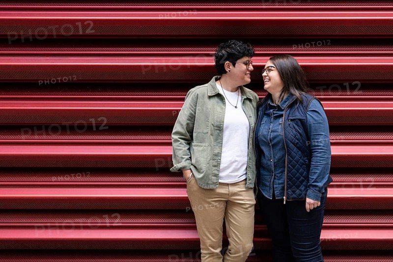 A joyful lesbian couple shares a loving moment in front of a red urban backdrop. The casual attire and genuine smiles reflect their affection and happiness