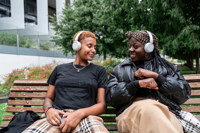 A joyful LGBT multiethnic couple listens to music with headphones, sitting on a park bench. They share smiles and connection in a serene outdoor setting, surrounded by greenery
