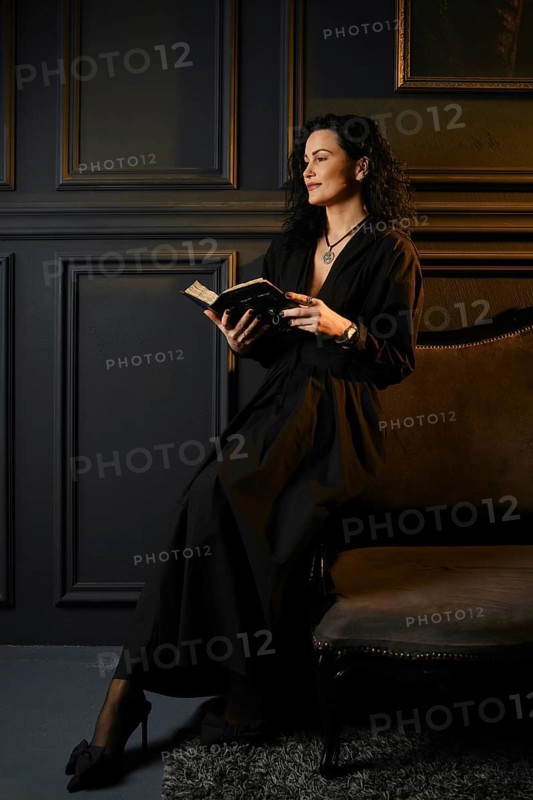 A woman sits on a chair in a dark room and reads a book. The walls are dark with paneling, and there is a piece of art in the background. The woman has long hair and wears a simple outfit