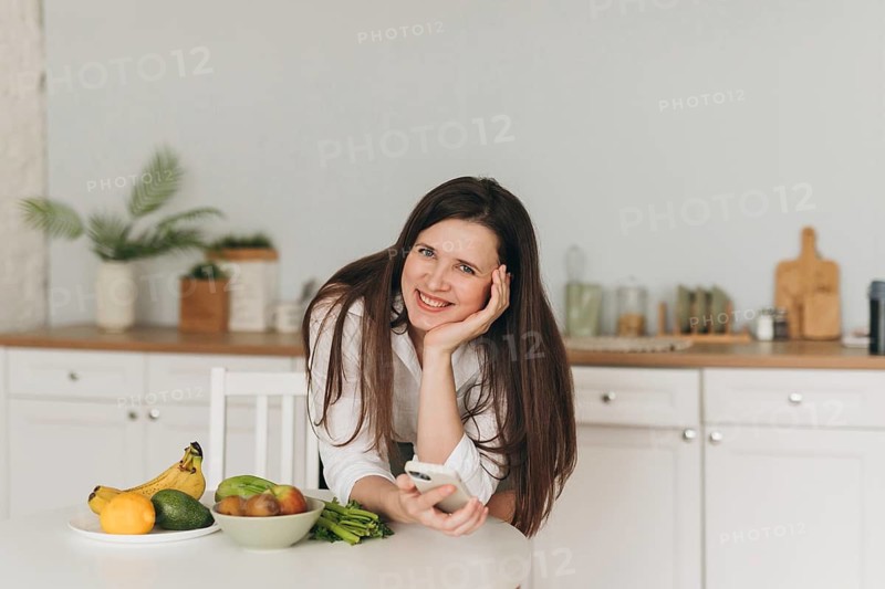A nutritionist smiles in a modern kitchen, surrounded by fresh fruits like bananas, avocados, and grapefruits. The bright setting promotes healthy living and balanced diet concepts