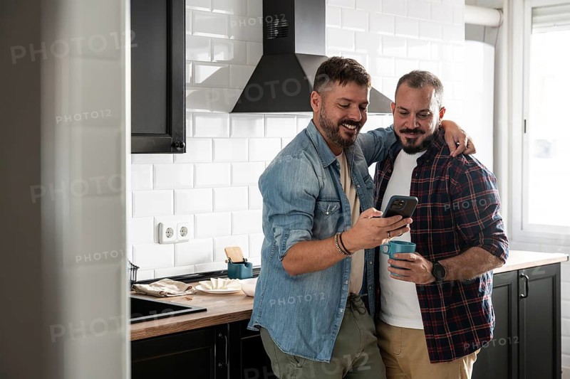 A gay couple shares a tender moment in their modern kitchen. One holds a phone while the other embraces him, both smiling warmly and enjoying each other's company