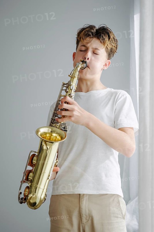 A young boy in a white shirt skillfully plays a saxophone, eyes closed in concentration. The soft lighting and muted background create an intimate musical setting