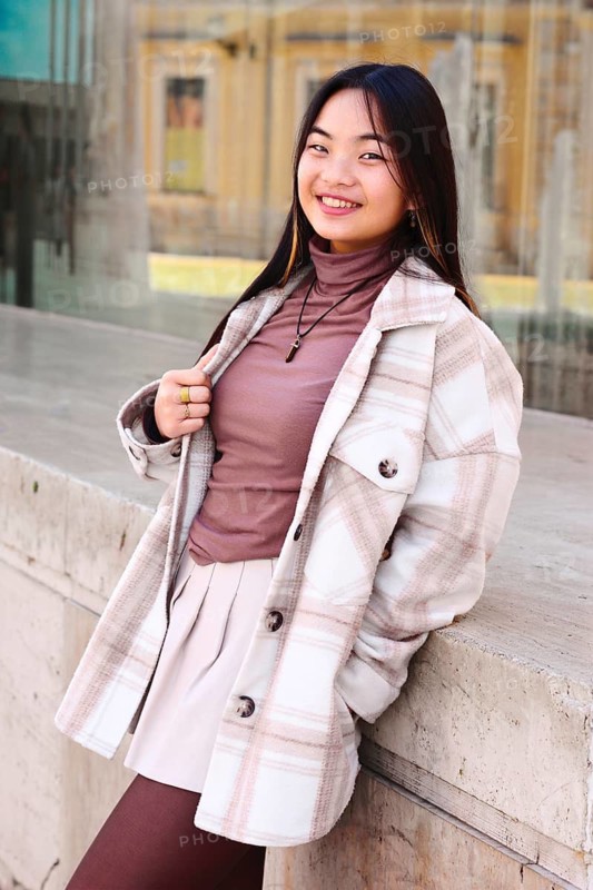 Young woman joyfully standing outdoors in a stylish autumn ensemble, including a plaid coat, turtleneck, and skirt. Her vibrant smile exudes warmth and confidence
