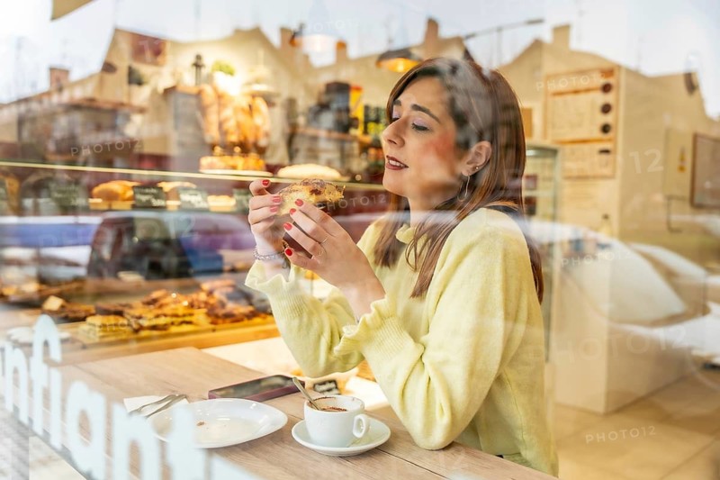 Woman savoring a flaky pastry and steaming coffee at a sunlit cafe window, enjoying a relaxed morning breakfast moment of indulgence and quiet contemplation