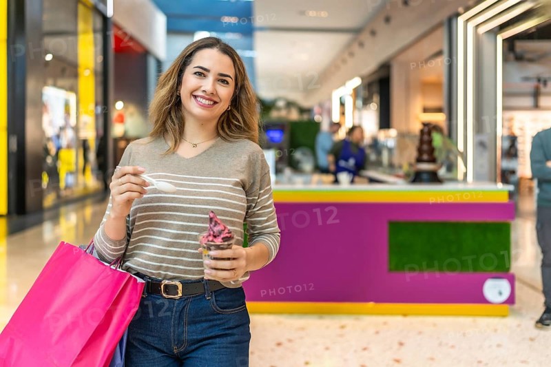 Happy young woman smiling at camera while holding shopping bags and eating ice cream in a bright busy mall, enjoying leisure time, retail therapy and a sweet treat