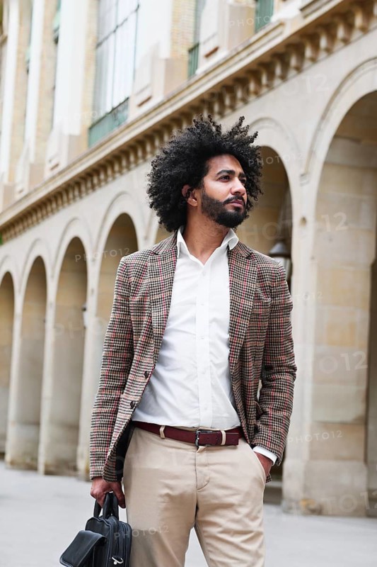 A mixed-race insurance salesman with afro hair stands confidently outdoors, dressed in a plaid blazer and white shirt. The background features classic stone arches on a building