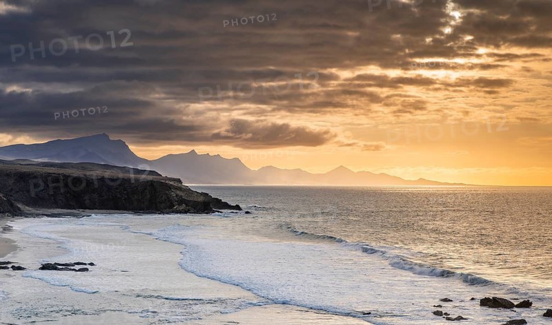 A stunning view of La Pared Beach in Fuerteventura at sunset. The golden light reflects on the ocean waves, while dramatic clouds create a breathtaking coastal landscape