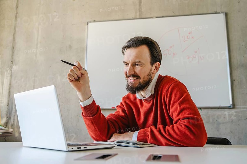 A man in a red sweater smiles while holding a pen and engaging with his laptop in a casual office setting, with a whiteboard in the background. Perfect for business or educational themes