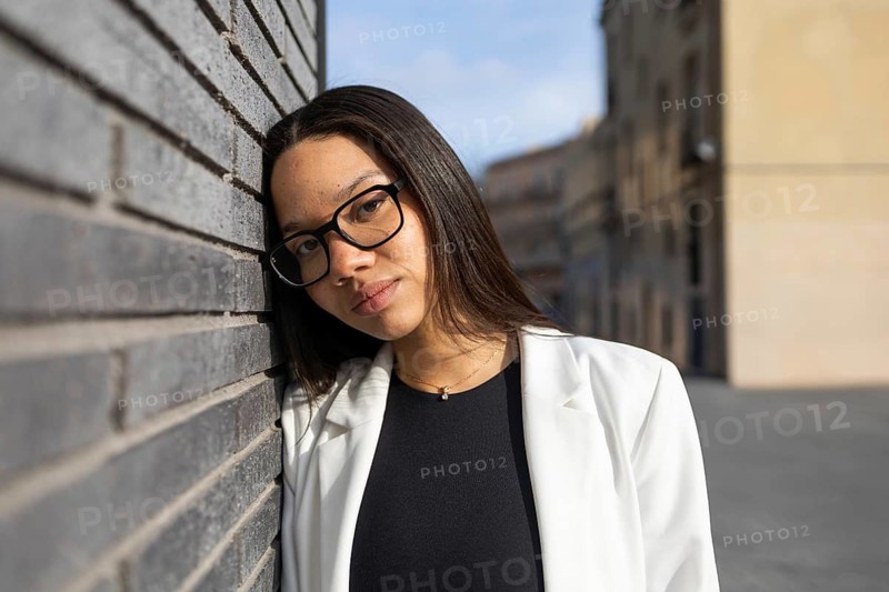 A professional woman with glasses leans against a brick wall. Her white blazer contrasts with the urban background, highlighting her confident business lifestyle