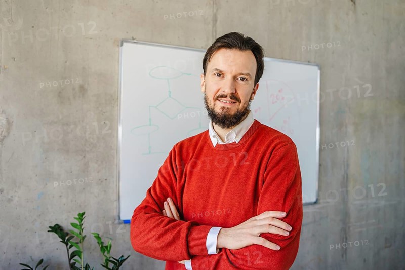 A man in a red sweater stands with arms crossed in front of a whiteboard in a modern office setting, exuding professionalism and leadership