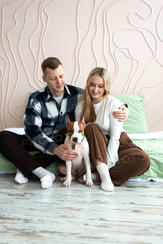 A young man and woman are seated on the floor at home, smiling joyfully as they interact with their white and tan dog. They display affectionate body language, emphasizing a comfortable domestic life