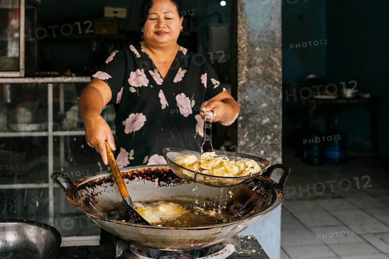Woman expertly fries in hot oil using a large wok. The scene captures a traditional cooking technique in a rustic kitchen, showcasing the culinary skill of Bali